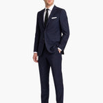 A man modeling a navy blue wool two-piece suit with a white dress shirt, black tie, and white pocket square, standing against a plain white background.
